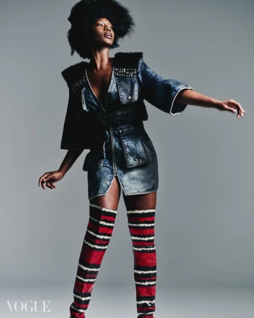Fashion editorial shot of a Black woman in denim dress, fur accents, and thigh-high striped boots posing against a neutral gray background.