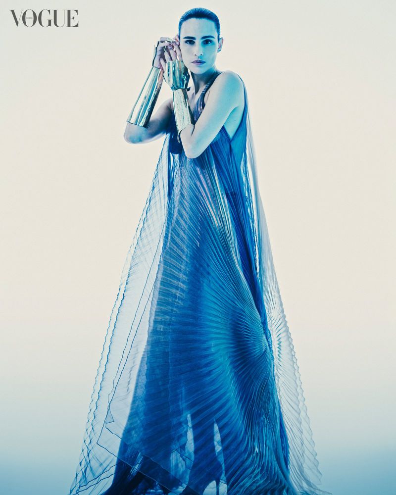 Editorial fashion shot of a woman in a blue dress and gold arm cuffs posing in a bright, neutral studio, featuring Vogue magazine logo.