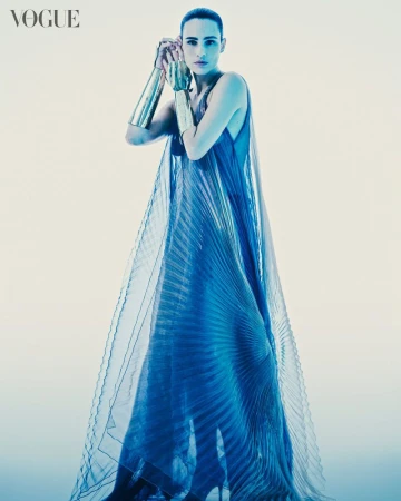 Editorial fashion shot of a woman in a blue dress and gold arm cuffs posing in a bright, neutral studio, featuring Vogue magazine logo.