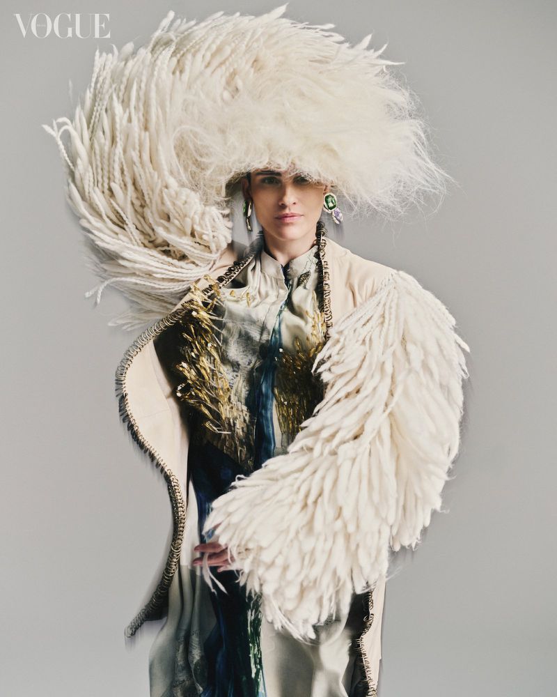 Fashion editorial shot of a woman wearing an extravagant feathered hat and coat in a studio setting, highlighting avant-garde design and texture.