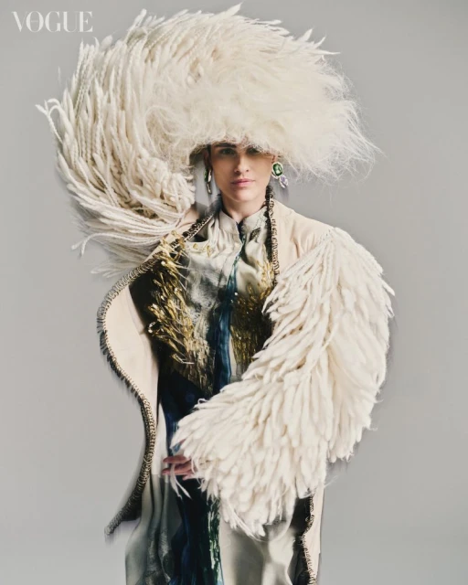 Fashion editorial shot of a woman wearing an extravagant feathered hat and coat in a studio setting, highlighting avant-garde design and texture.