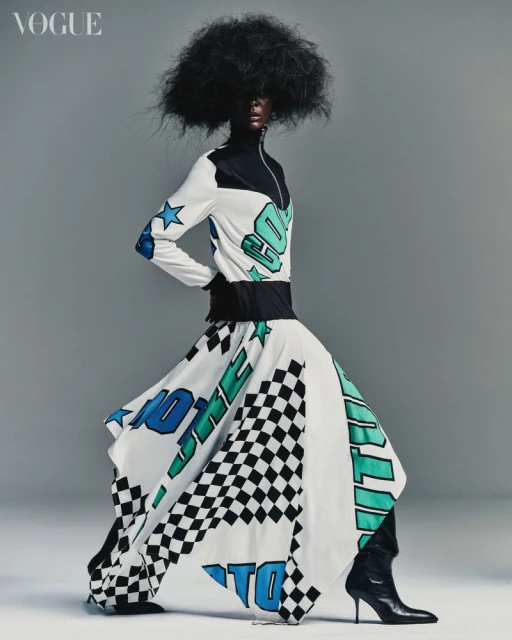 Fashion shot of a woman with dark skin and a large afro wearing a colorful printed dress, black heeled boots, posing in a photo studio.