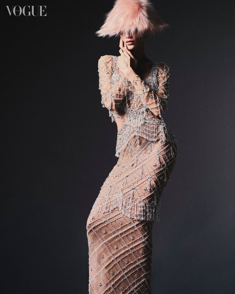 Fashion shot of a female model in an elaborate beaded dress and pink feather hat, posing against a dark gray background for Vogue.