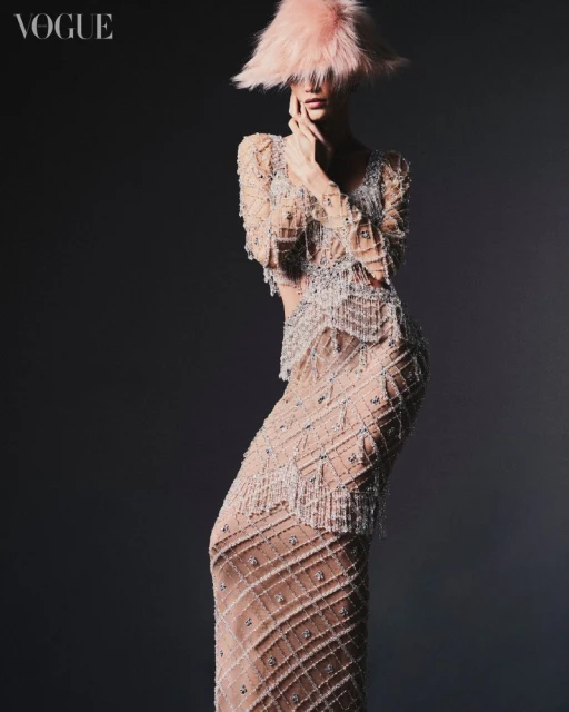 Fashion shot of a female model in an elaborate beaded dress and pink feather hat, posing against a dark gray background for Vogue.
