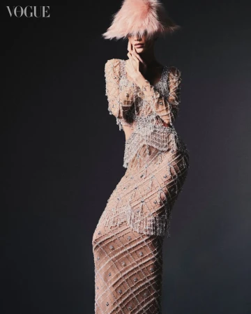 Fashion shot of a female model in an elaborate beaded dress and pink feather hat, posing against a dark gray background for Vogue.