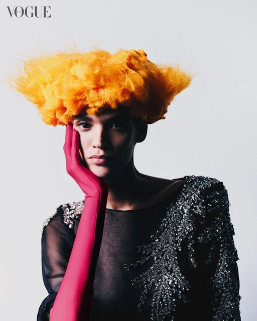 Fashion portrait of a female model wearing an embellished black dress, orange wig, and long pink glove, posing in a photo studio.