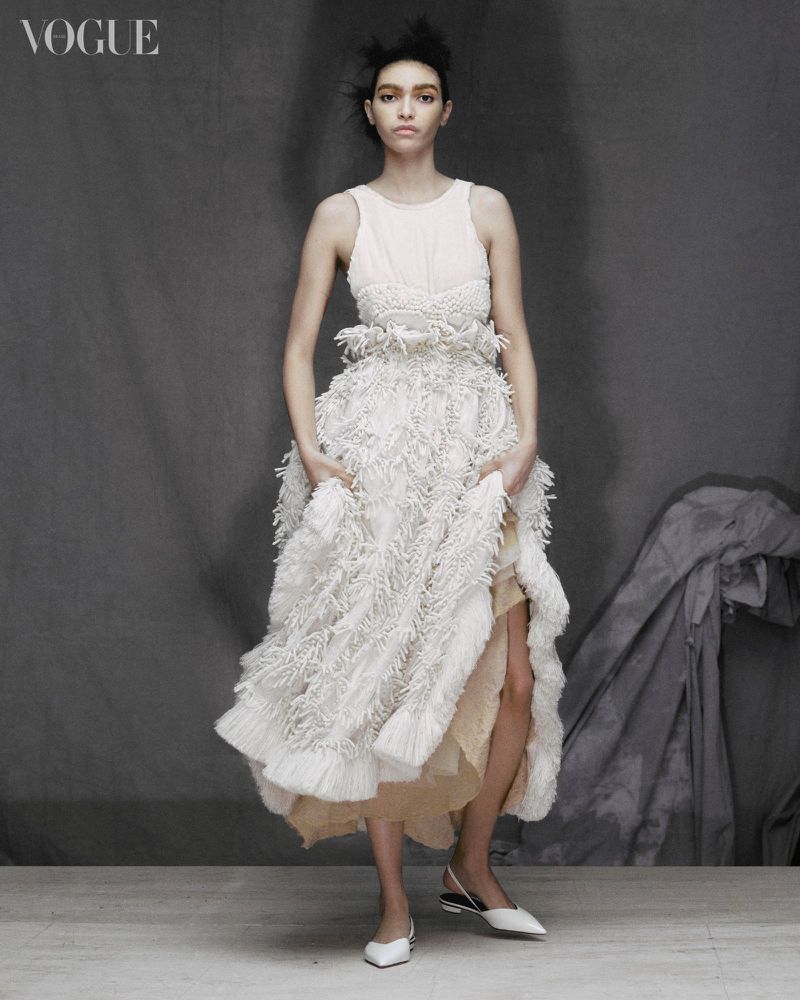 Full-length portrait of a young woman wearing a white textured dress, posing in front of a grey background inside a photo studio.