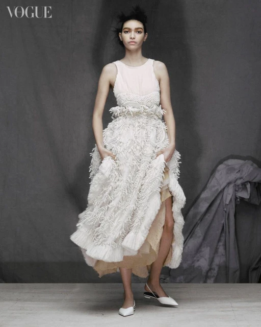 Full-length portrait of a young woman wearing a white textured dress, posing in front of a grey background inside a photo studio.