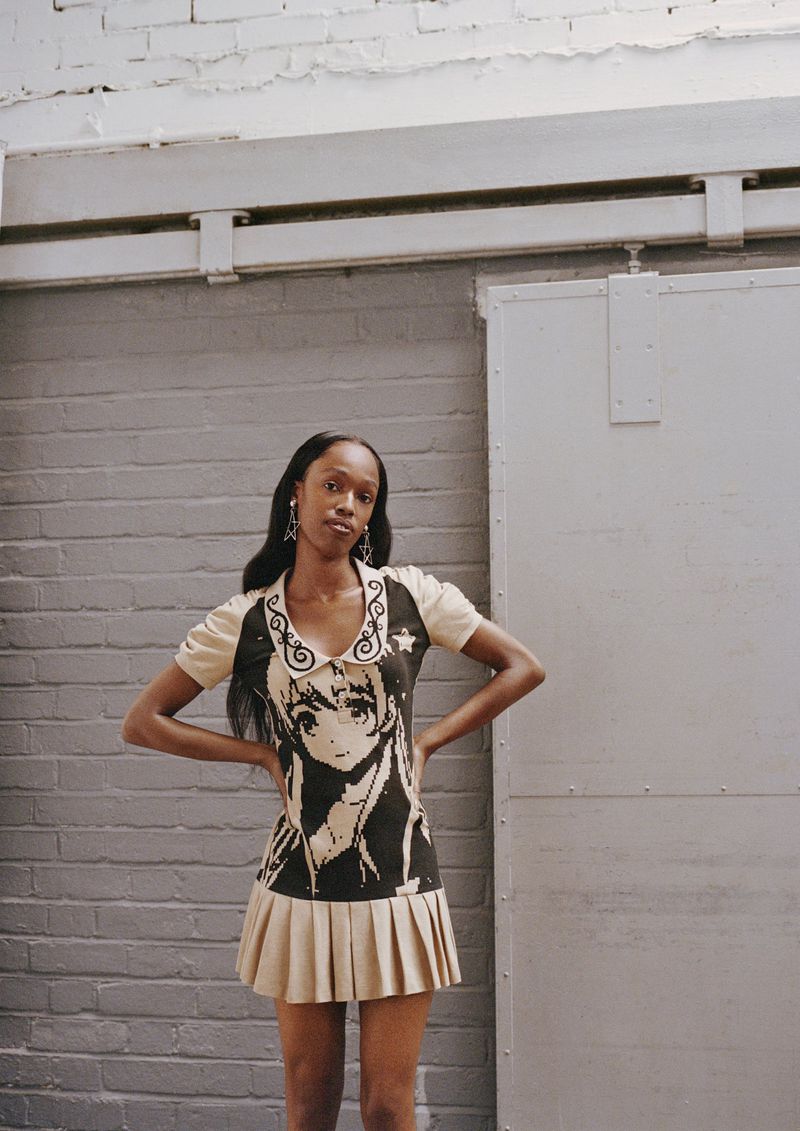 A fashionable young woman poses in a knit dress with an anime character graphic, set against a brick wall in a brightly lit indoor space.