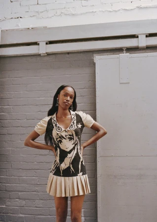 A fashionable young woman poses in a knit dress with an anime character graphic, set against a brick wall in a brightly lit indoor space.