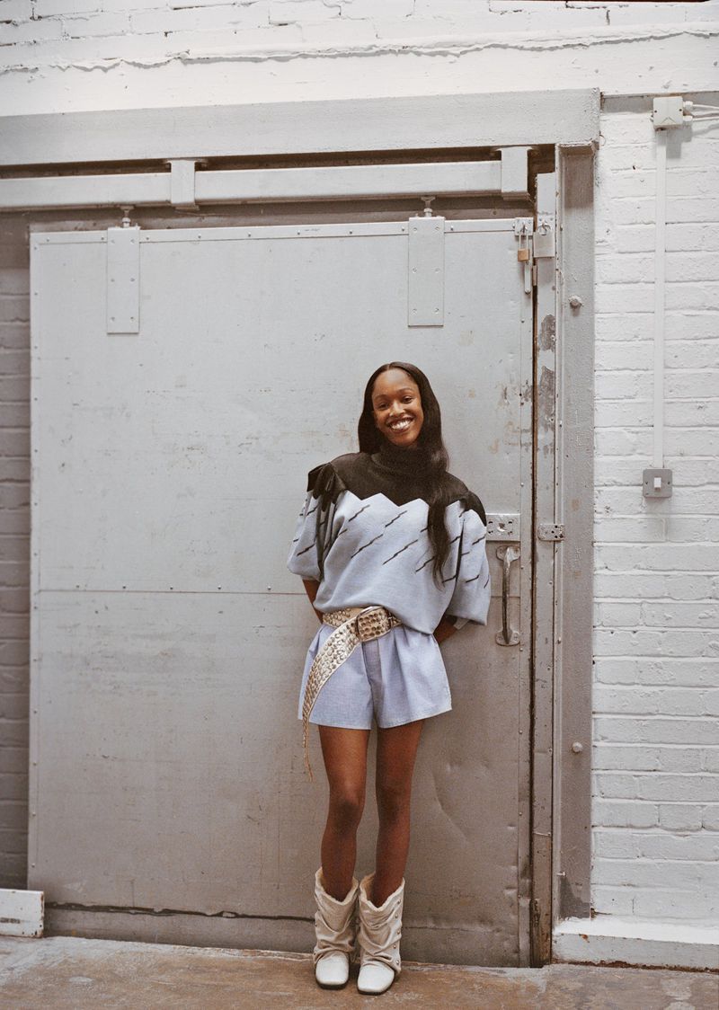 A smiling African American woman poses against a gray loading dock door, wearing a fashionable blue shorts suit and cowboy boots.