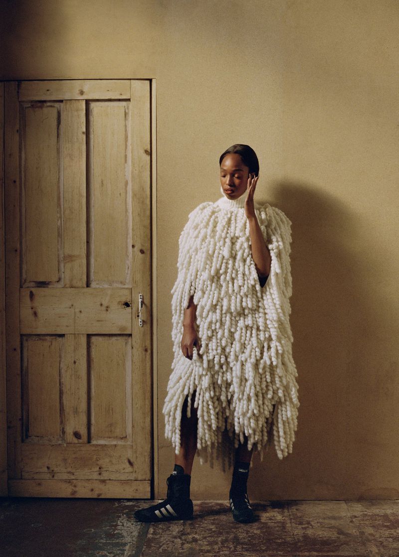 A woman in a cream-colored, fringed cape stands in front of a wooden door against a neutral wall, wearing black Adidas wrestling shoes.