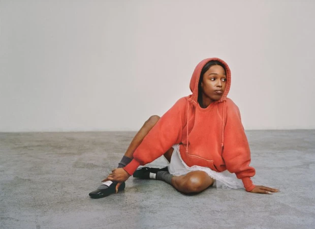 Young woman in red hoodie sits on grey floor in studio, wearing skirt, socks and loafers, gazing left with neutral expression and soft lighting.