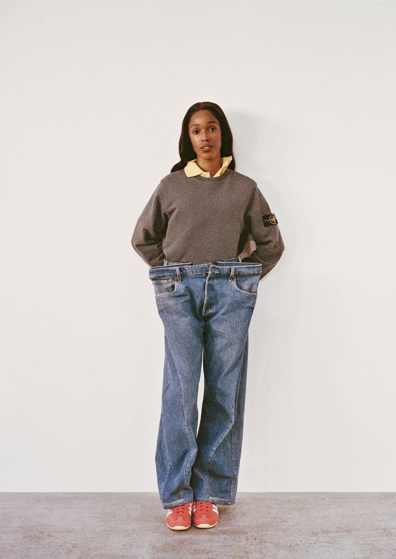 Fashion shot of a woman wearing a crew neck sweater and jeans standing in front of a white wall, showcasing a modern aesthetic.