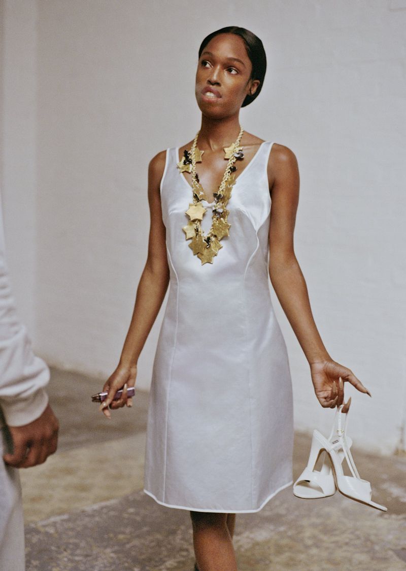 A fashionable young woman in a white dress and star necklace holds heels, posing in a minimalist studio with concrete floors.
