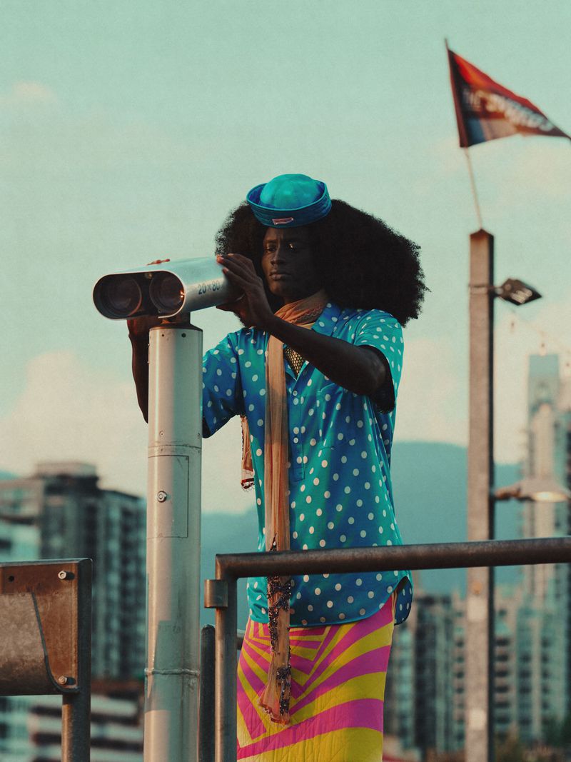An adult male looking through binoculars in an urban setting, wearing a blue polka dot shirt and colorful striped skirt with city backdrop.