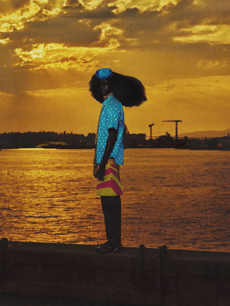 Person in retro fashion stands near a body of water at sunset. The individual wears a polka dot shirt, hat, and striped shorts.