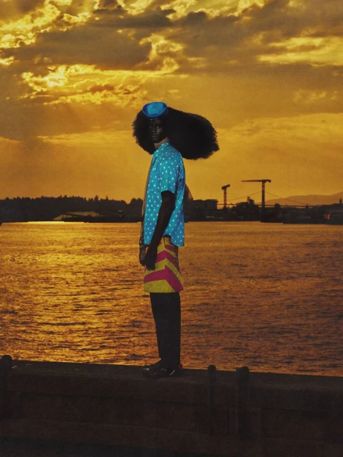 Person in retro fashion stands near a body of water at sunset. The individual wears a polka dot shirt, hat, and striped shorts.