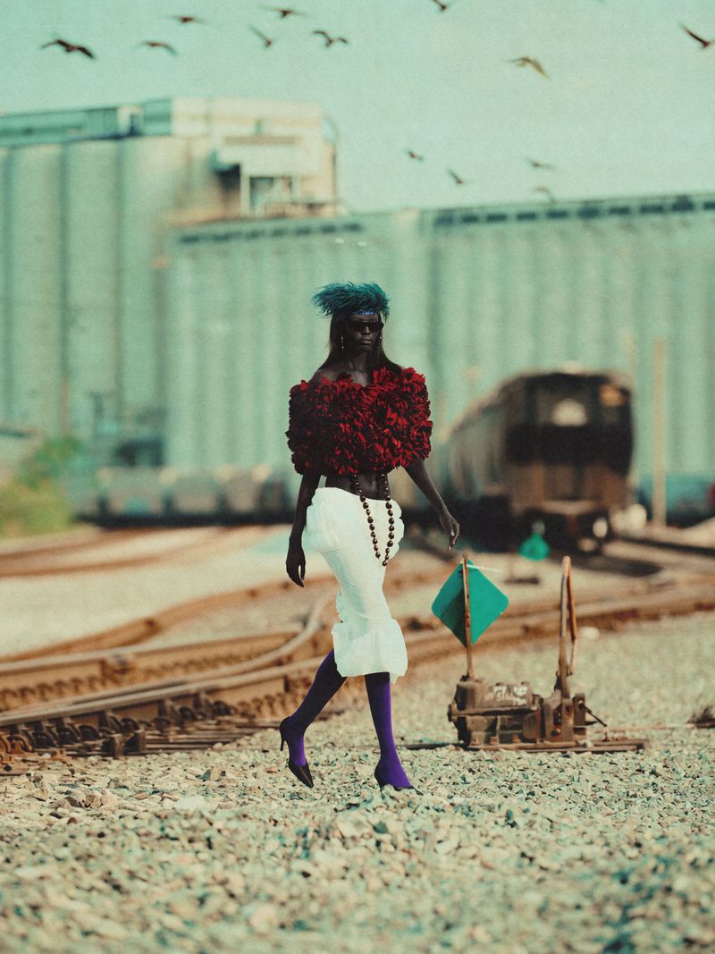 Fashionable woman in red top and white skirt walks along railroad tracks with industrial buildings and a flock of birds in the distance.