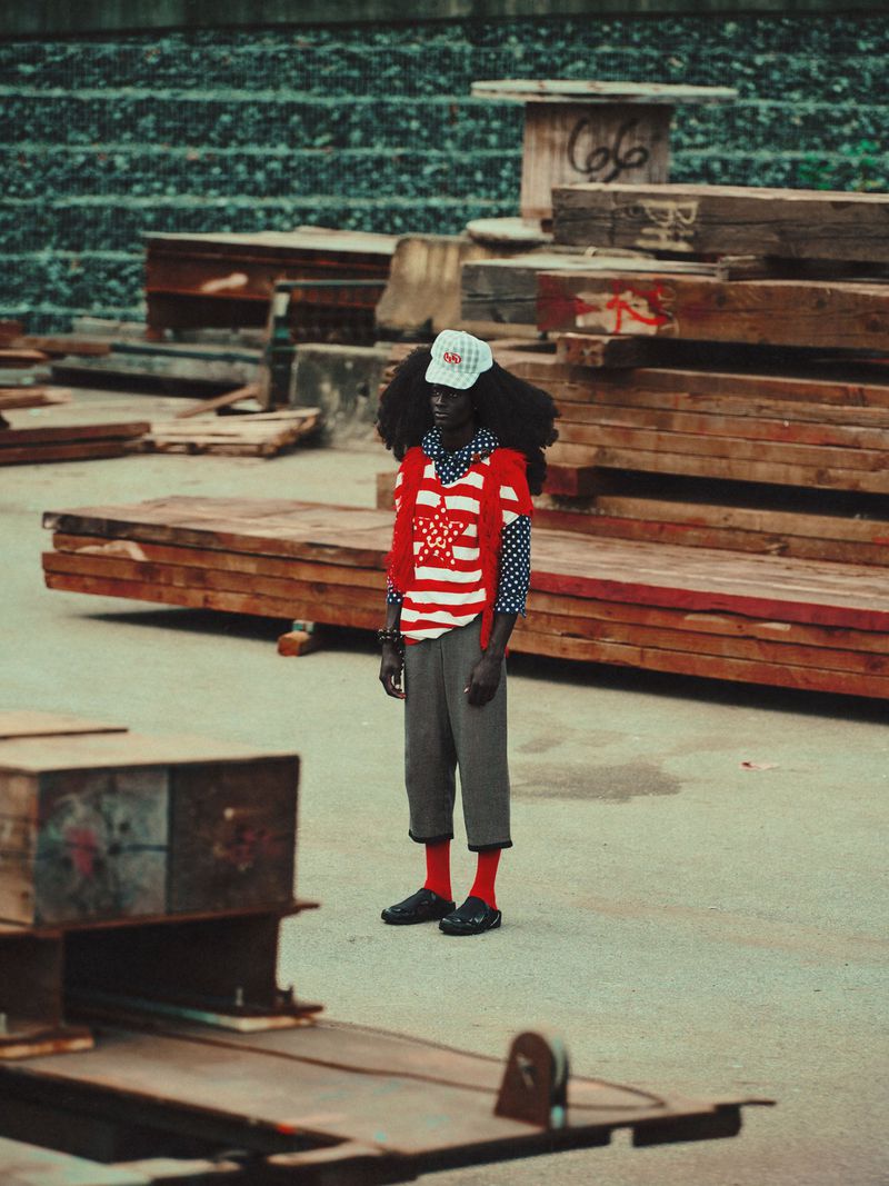 Man with afro wearing stars and stripes stands outdoors amongst wood piles and gray concrete under overcast daylight in a medium shot.