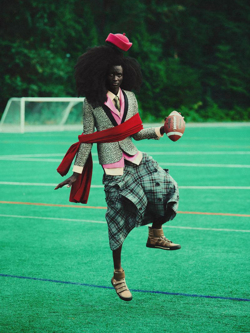 Fashionable adult posing on green sports field wearing suit jacket, kilt, and red cap holding football against a green tree background.