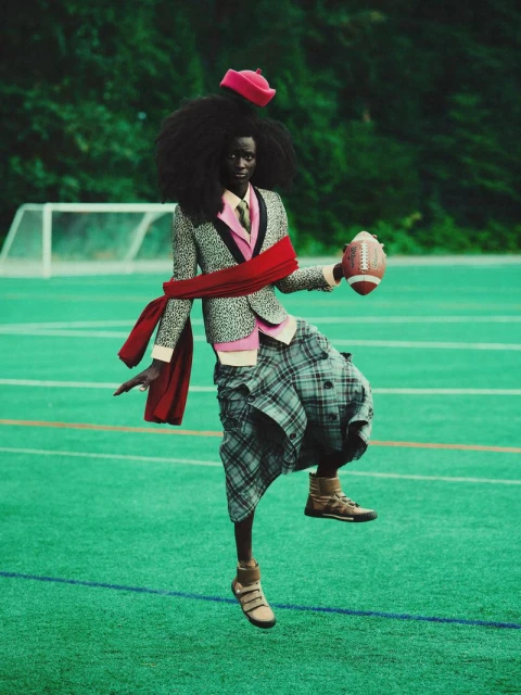 Fashionable adult posing on green sports field wearing suit jacket, kilt, and red cap holding football against a green tree background.