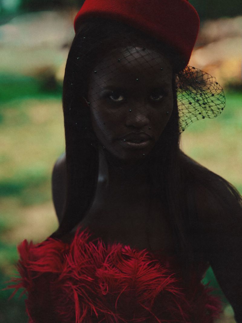 Close-up portrait of a stylish dark-skinned woman with a serious facial expression, wearing a red hat and feathered top in a nature setting.