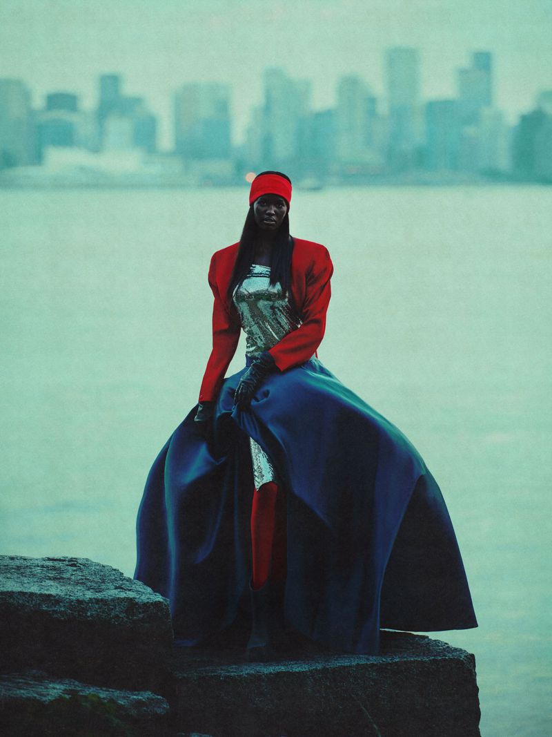 Woman in a shiny silver top and red and blue dress sitting on rocks near a city skyline and a large expanse of water.