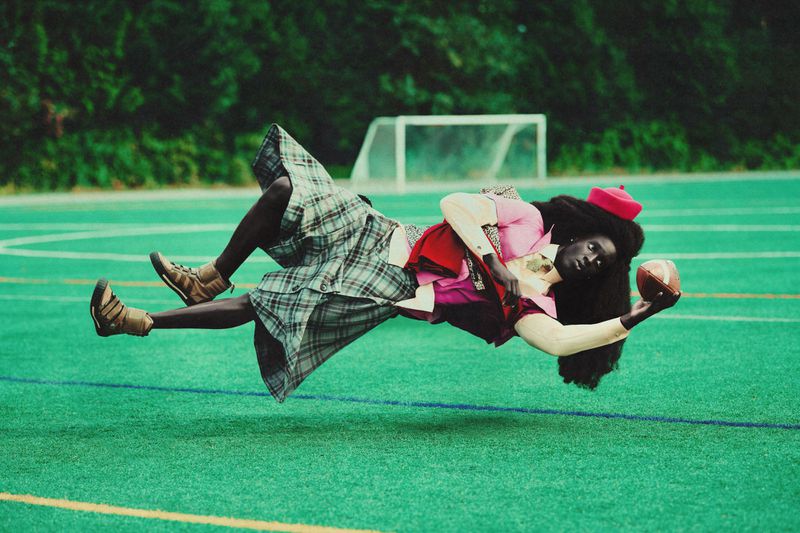 Young woman with long dark hair, wearing a pink top, plaid skirt, and red beret, appears to be floating with a football in her hand.