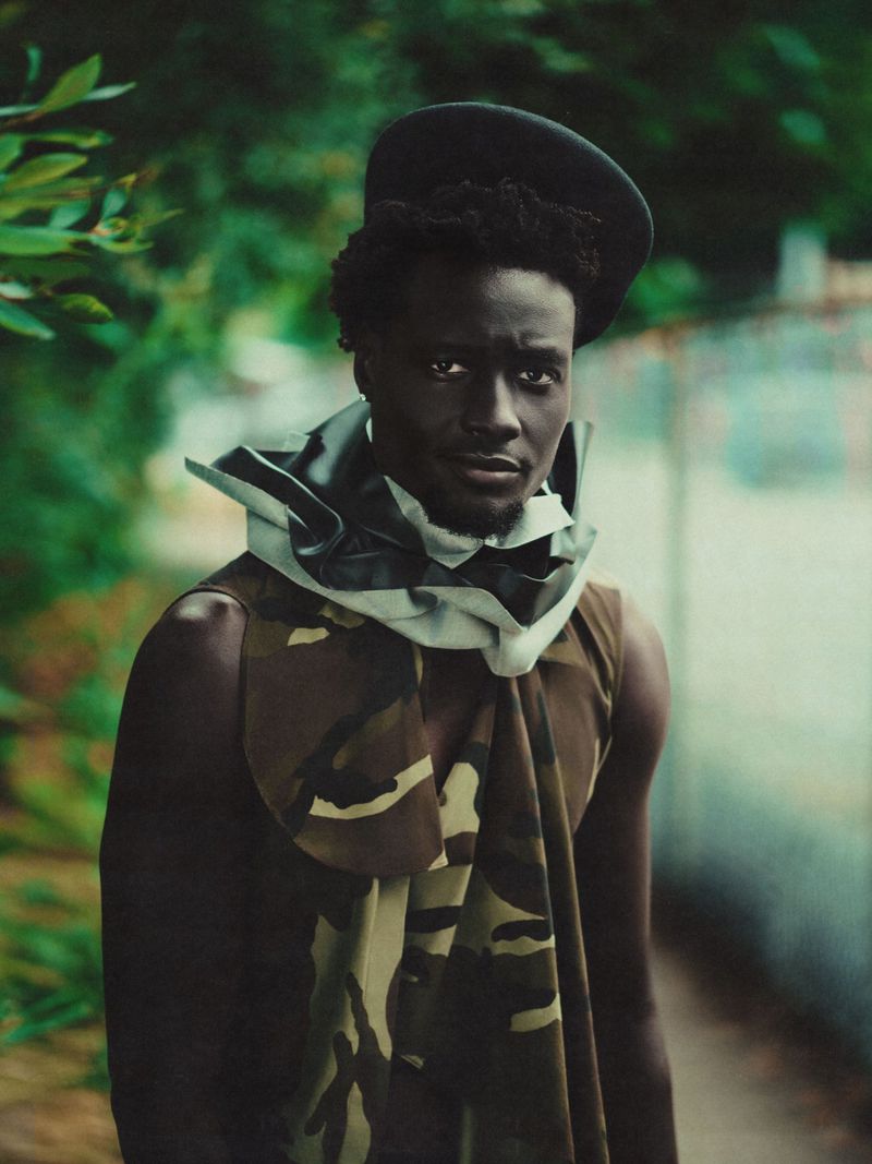 Stylish Black male model in camouflage fashion, headwear, posing outdoors with a serious facial expression, lush green background.