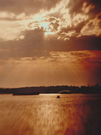 Scenic view of sunlight breaking through clouds above a body of water with trees on the horizon, creating a tranquil, atmospheric landscape.