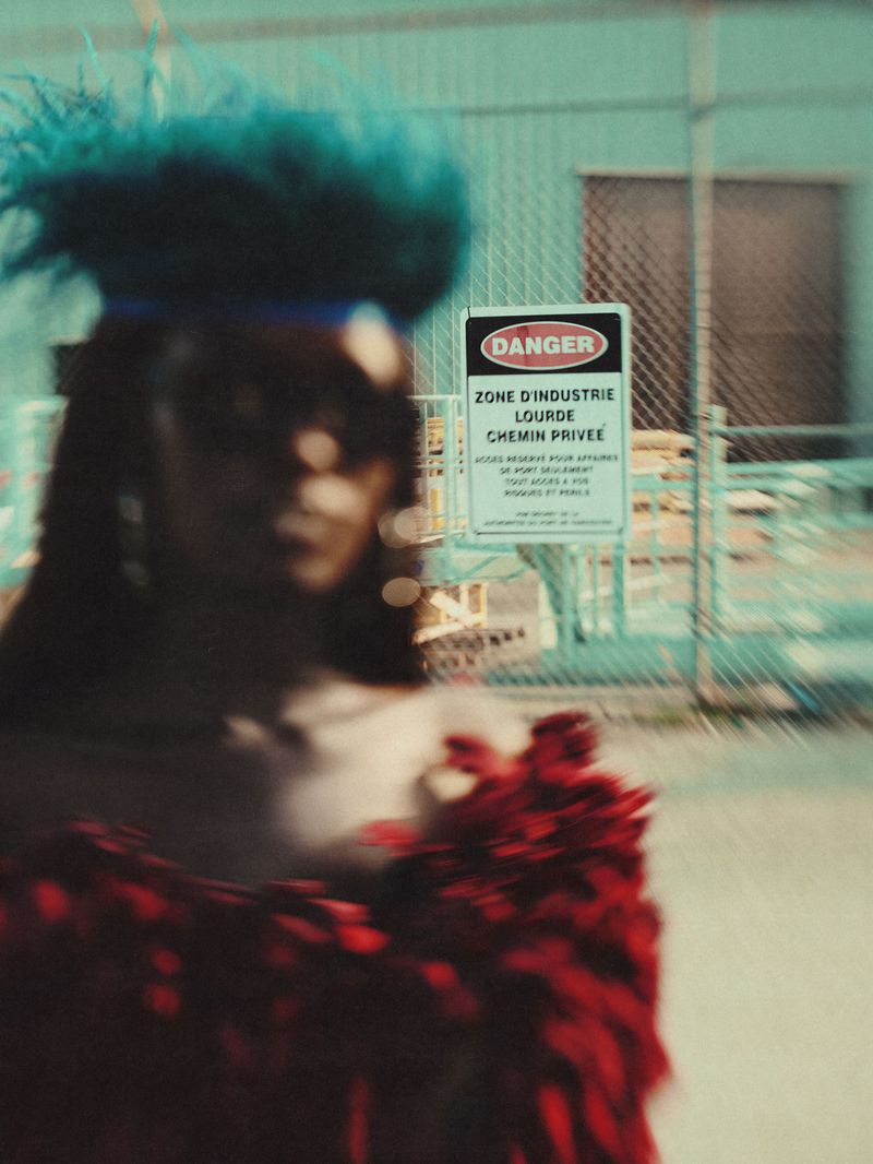 Model in motion wearing blue feathered headpiece and red feathered shawl with industrial site and danger sign in the blurred background.