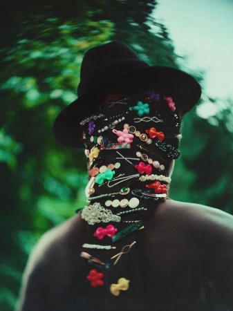 Person seen from behind wearing a black wide-brim hat and dark hair covered in colorful barrettes against blurred green foliage.
