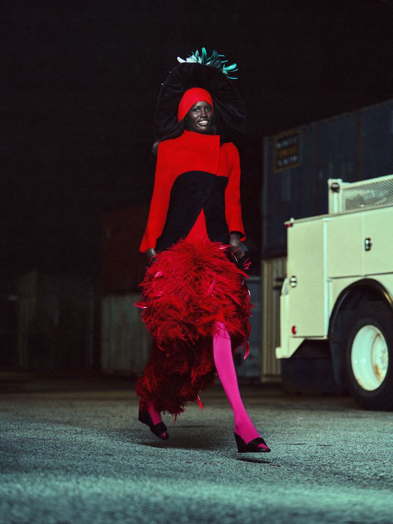 Elegant woman in a red and black dress with feather detailing, walking on a gray street with a large white truck in the background.