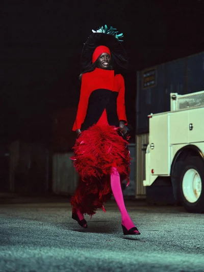 Model walking at night wearing an avant-garde red feathered skirt, red and black coat, large dark hat, and bright pink tights.