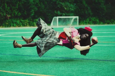 Model in plaid kilt and pink top floating horizontally over a green sports field holding a football for Galore Magazine.