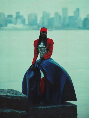 Fashion model in a red cropped jacket, metallic silver top, and wide blue skirt posing on coastal rocks with a skyline in the distance.