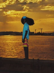 Model standing on a waterfront pier at sunset wearing a blue polka dot shirt and yellow patterned shorts, photographed by Gustavo Chams.