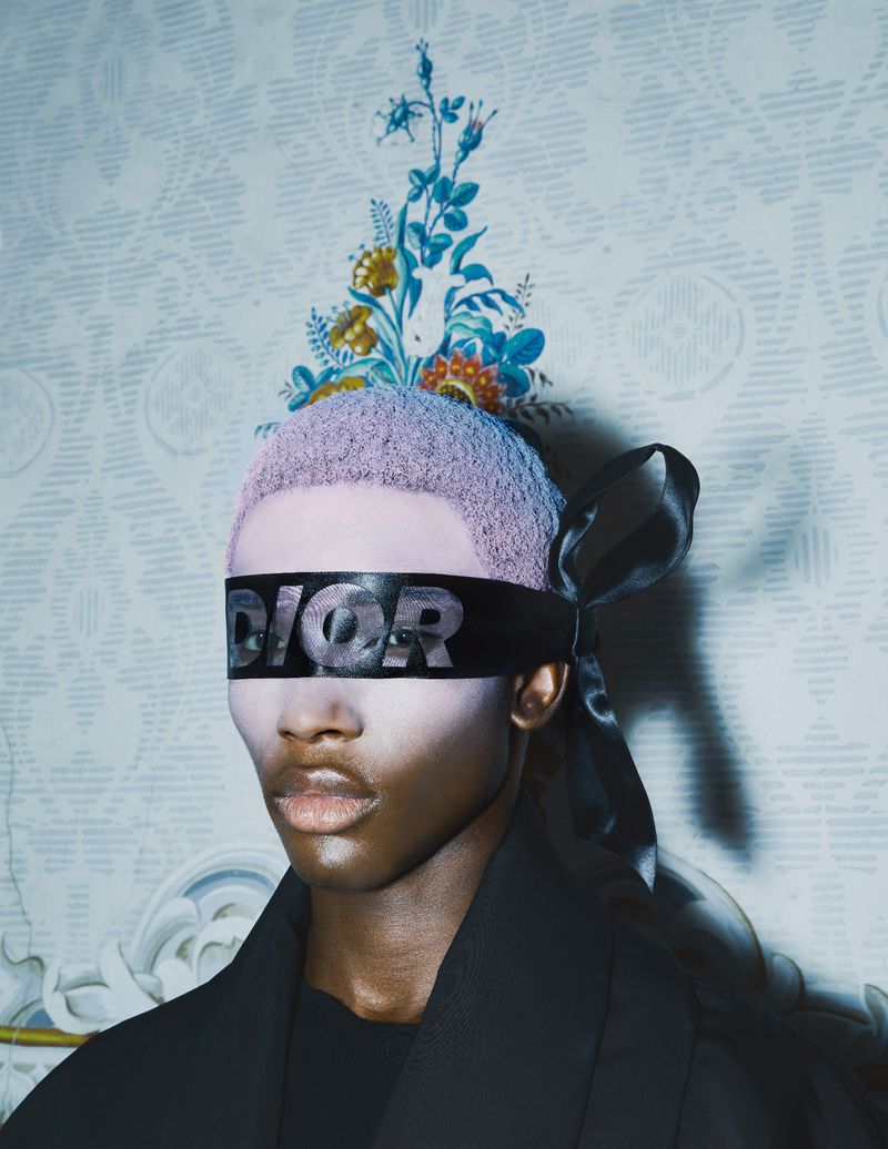 Fashion portrait of a non-binary person wearing a black Dior blindfold with lilac-colored hair in front of a patterned wallpaper.