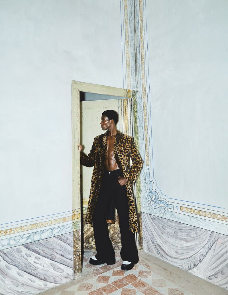 Fashionable Black male model in leopard coat stands in a room with painted walls and floors in a doorway looking left, editorial style.