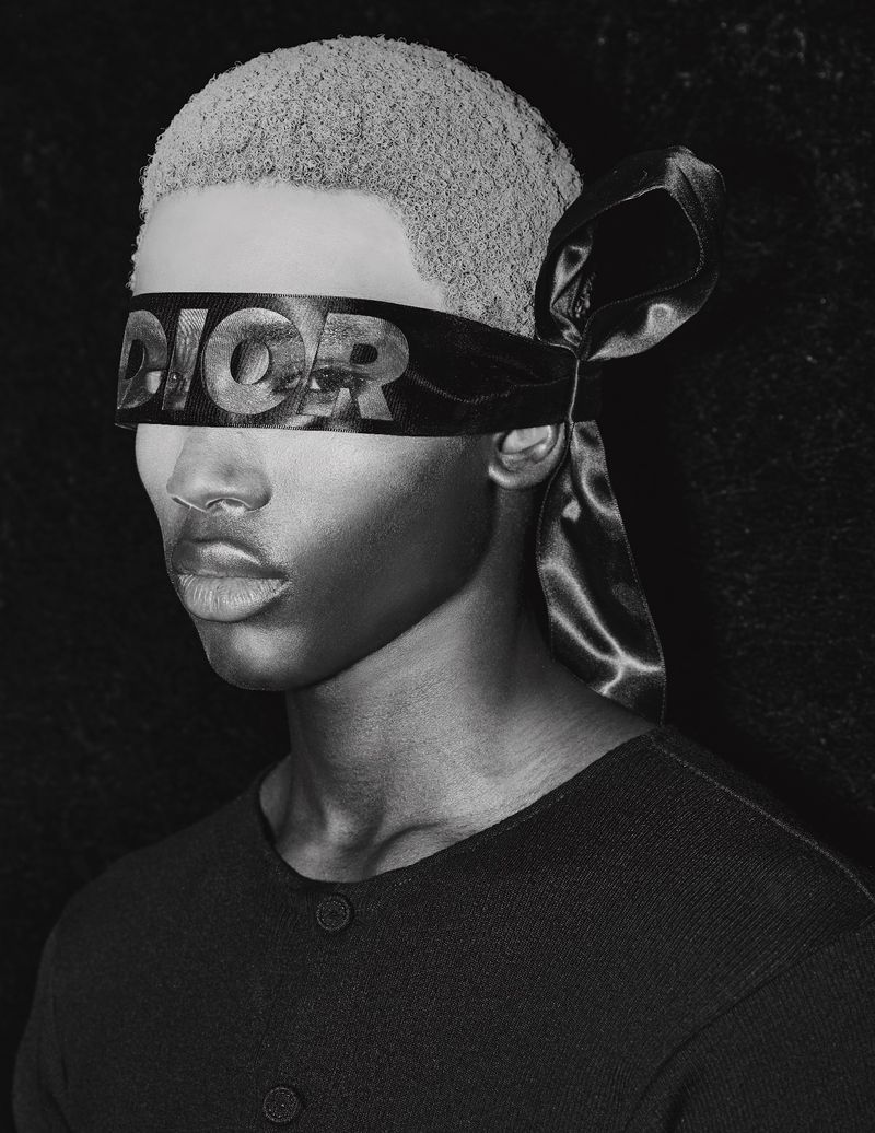 Monochrome portrait of a young adult with a Dior-branded blindfold, set against a dark background, shot with soft lighting.