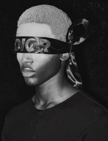 Monochrome portrait of a young adult with a Dior-branded blindfold, set against a dark background, shot with soft lighting.