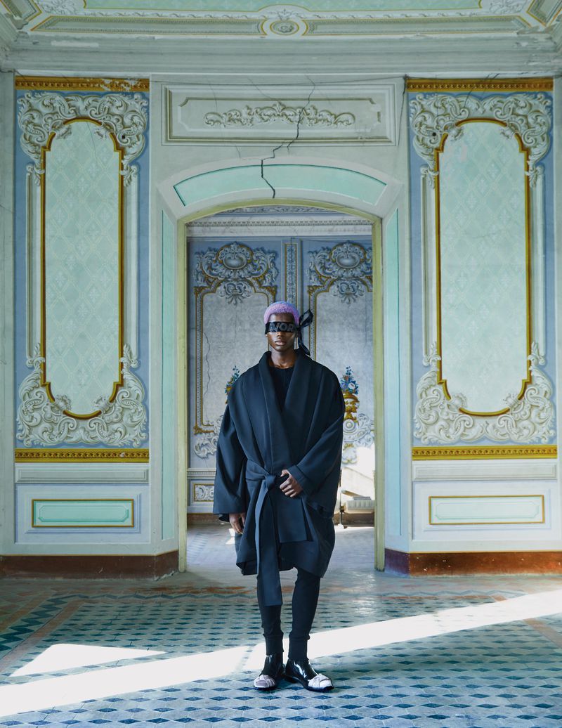 Person in a dark robe and headscarf standing in the doorway of an ornate, dilapidated room with patterned walls and floor tiles.
