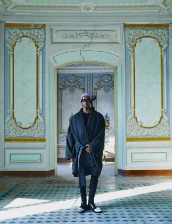 Person in a dark robe and headscarf standing in the doorway of an ornate, dilapidated room with patterned walls and floor tiles.