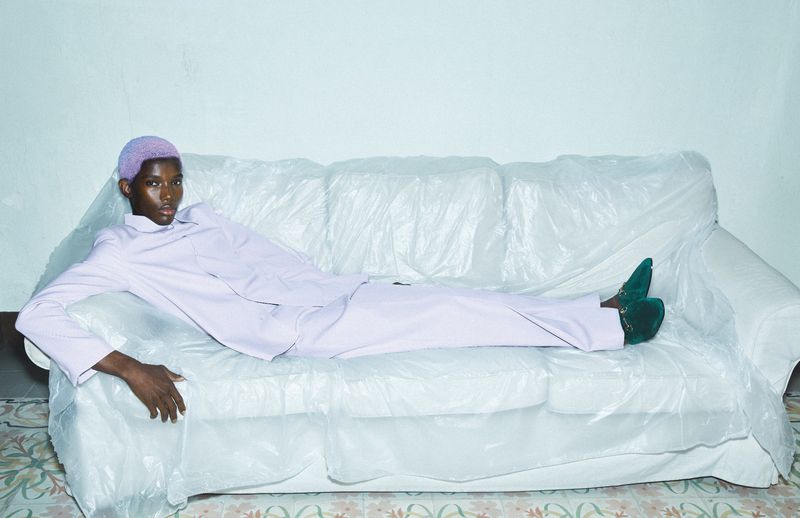 Stylish portrait of a black man wearing a lilac suit and vibrant shoes, reclining on a couch covered with translucent plastic sheeting.