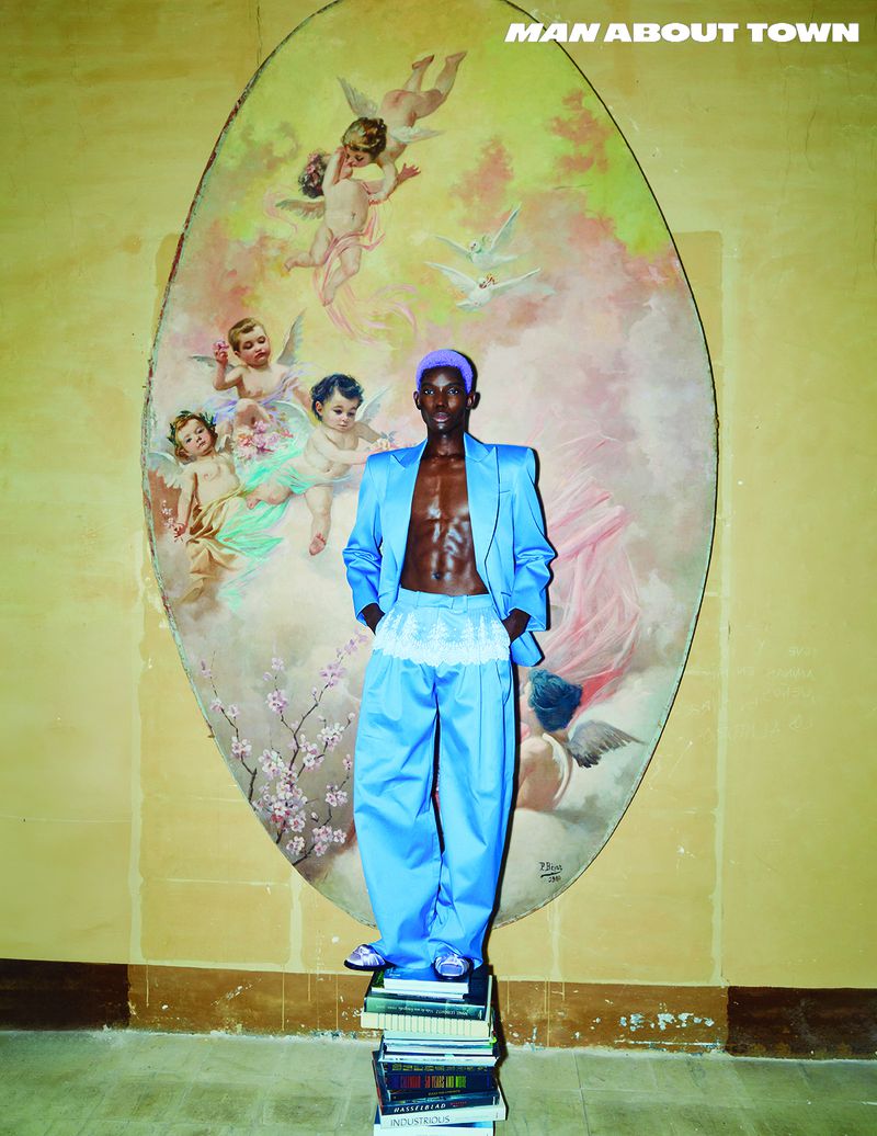 An African American man in a blue suit poses on a stack of books in front of a cherub mural with natural light illuminating the scene.