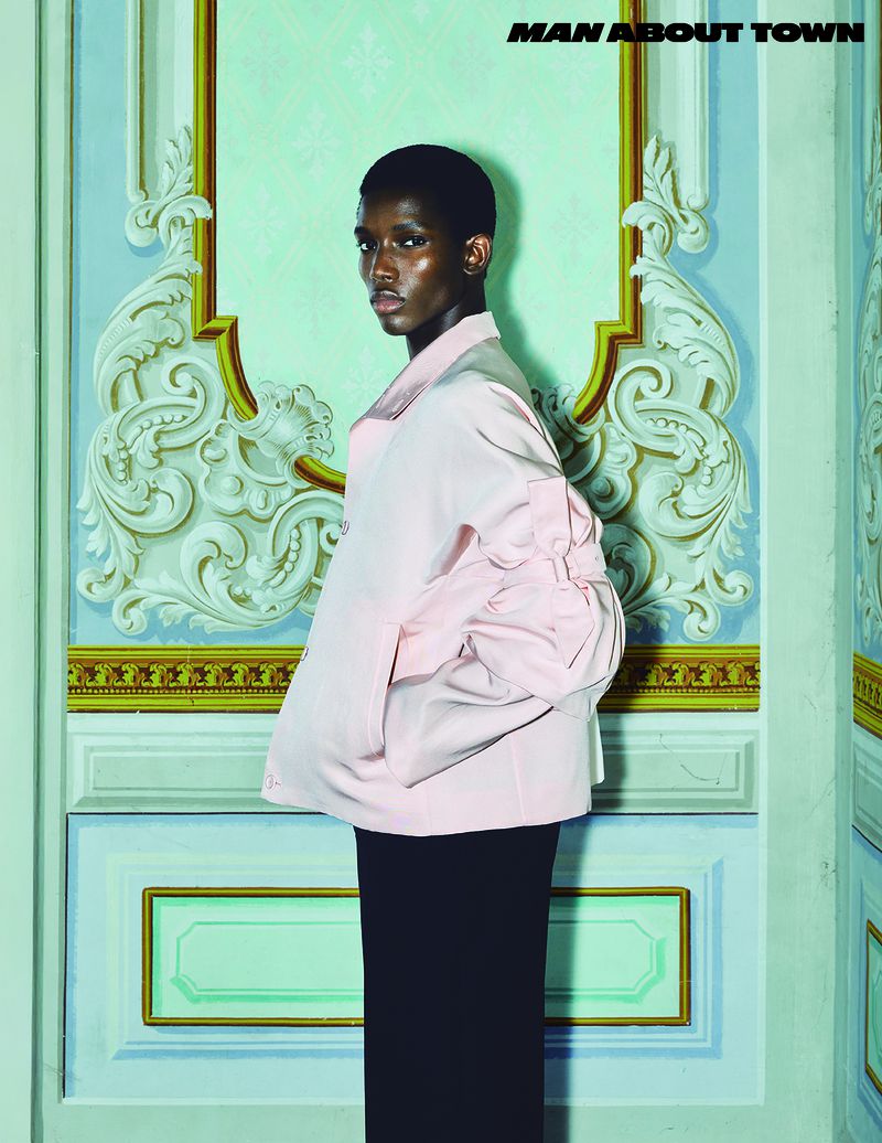 Fashion editorial portrait of a dark-skinned male model in a pink jacket and black pants, posing against an ornate wall.