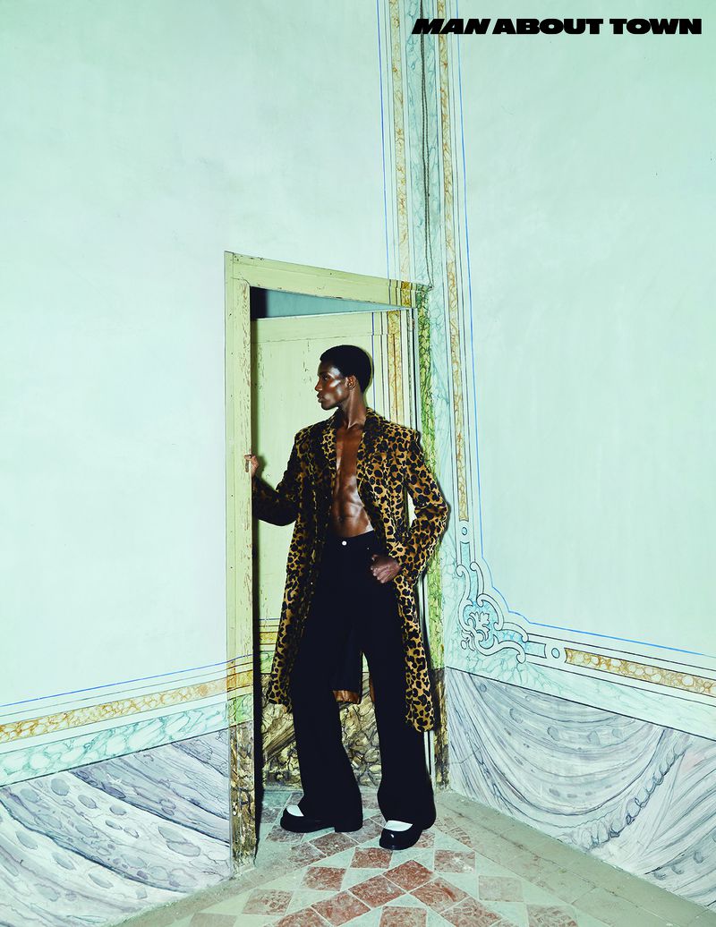 A Black male model poses in a cheetah-print coat and black pants in an ornately decorated room with a doorway behind him.