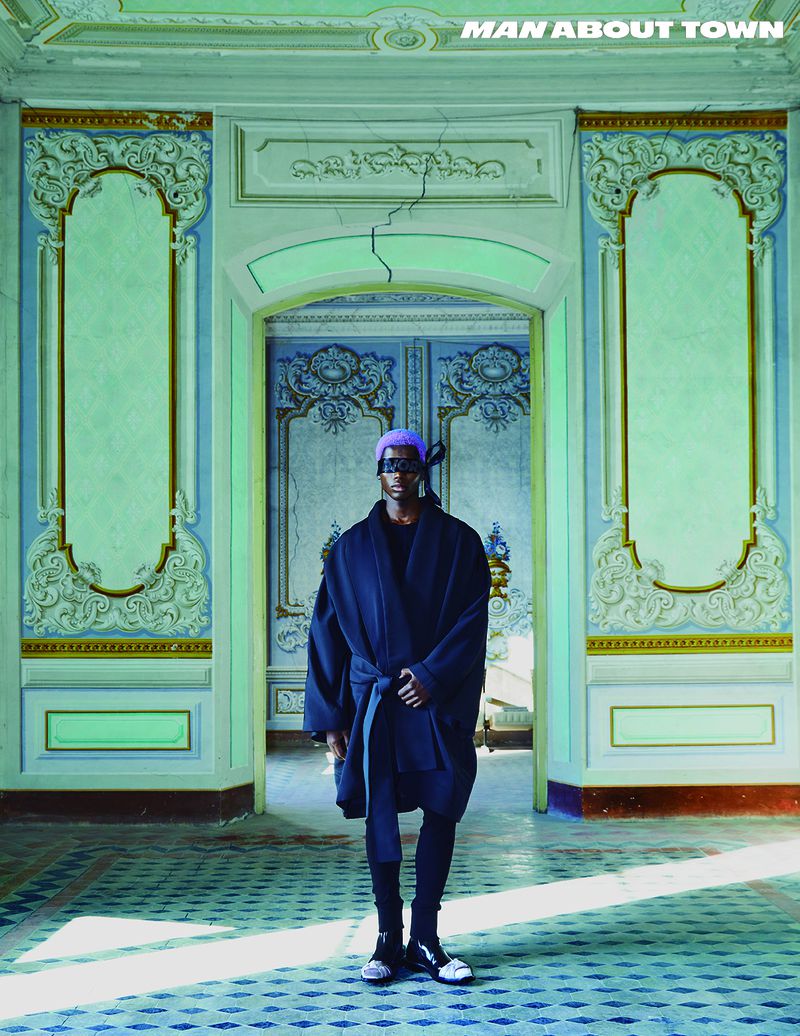 Editorial fashion shot of an adult male in a dark robe and purple head wrap standing in an ornate, rundown interior with a blue patterned floor.