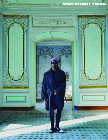 Editorial fashion shot of an adult male in a dark robe and purple head wrap standing in an ornate, rundown interior with a blue patterned floor.